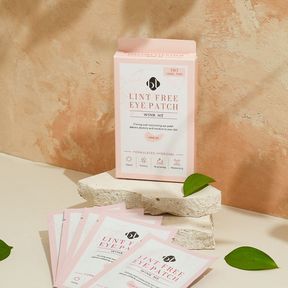 BL Lashes Lint Free Eye Patch ?œWink Me??hydrogel eye patch box displayed on a textured stone platform with several individual pink sachets spread in front. The packaging reads ?œLint Free Eye Patch Wink Me??and ?œFormulated Hydrogel,??with icons highlighting vitality, firming, brightening, and moisturizing benefits. The warm-toned background and green leaves create a natural beauty product composition designed for under-eye care during eyelash extension treatments.