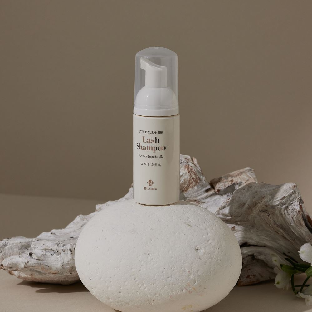 BL Lashes Lash Shampoo eyelid cleanser in a slim beige bottle with a foaming pump and transparent protective cap is placed upright on a smooth white stone, with the front label displaying “Eyelid Cleanser,” “Lash Shampoo,” “For Your Beautiful Life,” and “50 ml / 1.69 fl. oz.” while textured driftwood and small white flowers appear in the background, creating a natural and elegant beauty product composition that highlights a gentle lash cleansing product for eyelid hygiene and eyelash extension care.