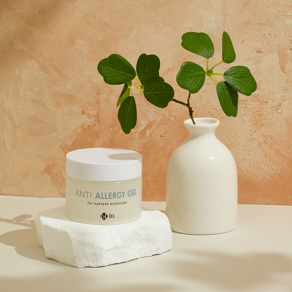 BL Lashes Anti Allergy Gel jar for eyelash extensions resting on a textured white stone, arranged with a white vase holding green leaves against a warm textured background.