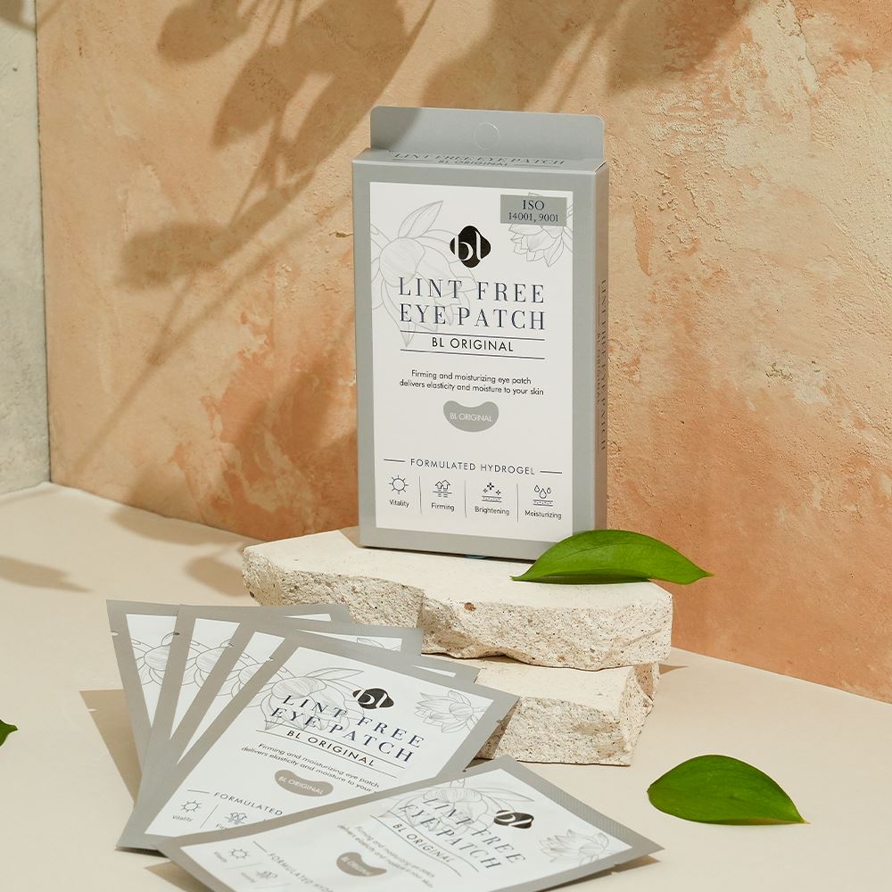 BL Lashes Lint Free Eye Patch ?œBL Original??hydrogel eye patch box displayed on stacked textured stone blocks with several individual gray sachets spread in front. The packaging reads ?œLint Free Eye Patch BL Original??and ?œFormulated Hydrogel,??with icons highlighting vitality, firming, brightening, and moisturizing benefits. Warm natural light, a textured beige background, and green leaves create a clean beauty product composition for under-eye care during eyelash extension treatments.