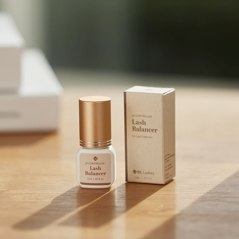 BL Lashes Lash Balancer pH Controller in a small frosted glass bottle with a metallic gold cap displayed beside its beige product box on a wooden surface. The label reads ?œpH Controller Lash Balancer??with the volume ?? ml / 0.17 fl. oz.??Warm natural light casts soft shadows, creating a lifestyle-style product presentation that highlights the compact professional solution designed for pH balancing during eyelash extension preparation.