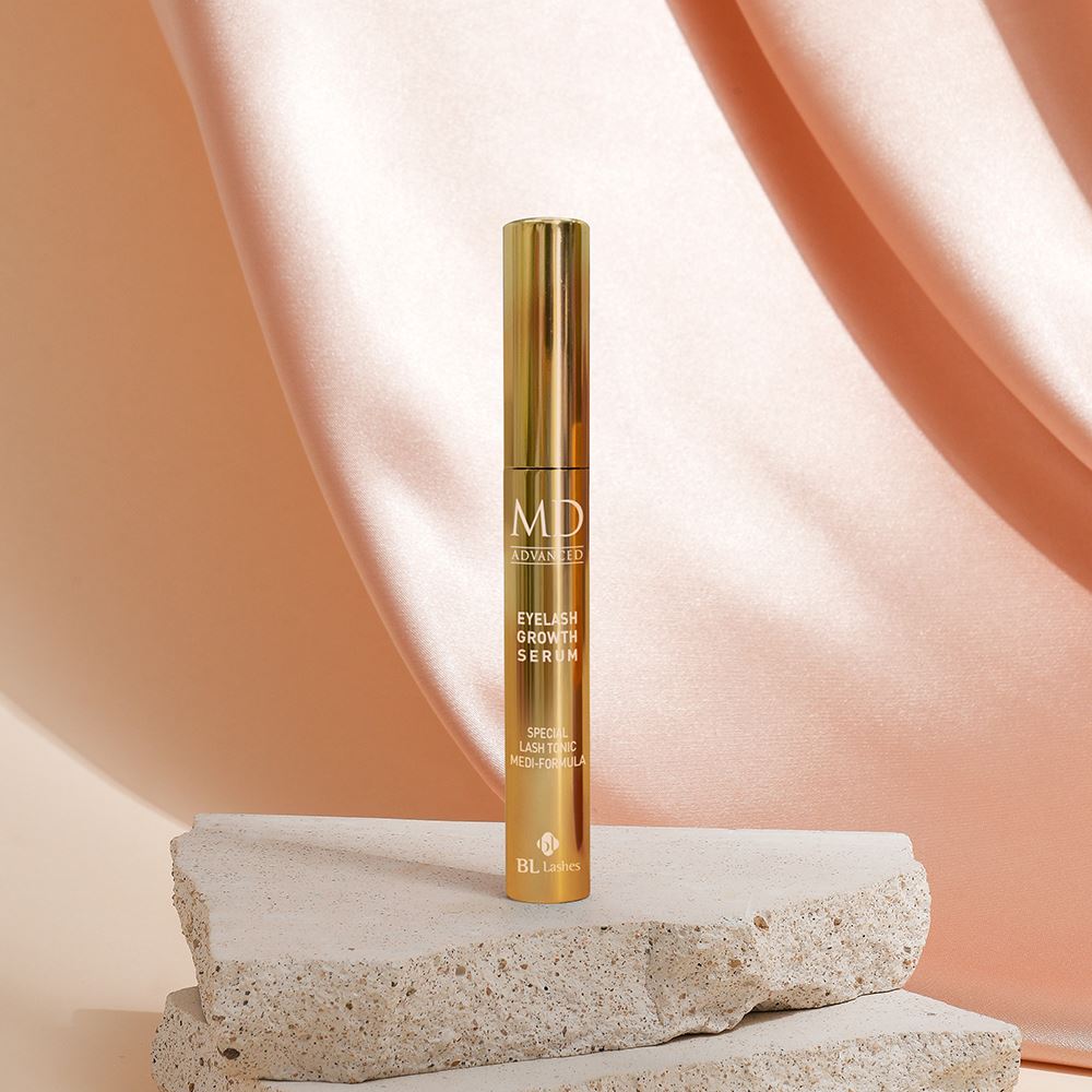 BL Lashes MD Advanced Eyelash Growth Serum gold tube displayed on textured stone blocks against a draped peach silk background.
