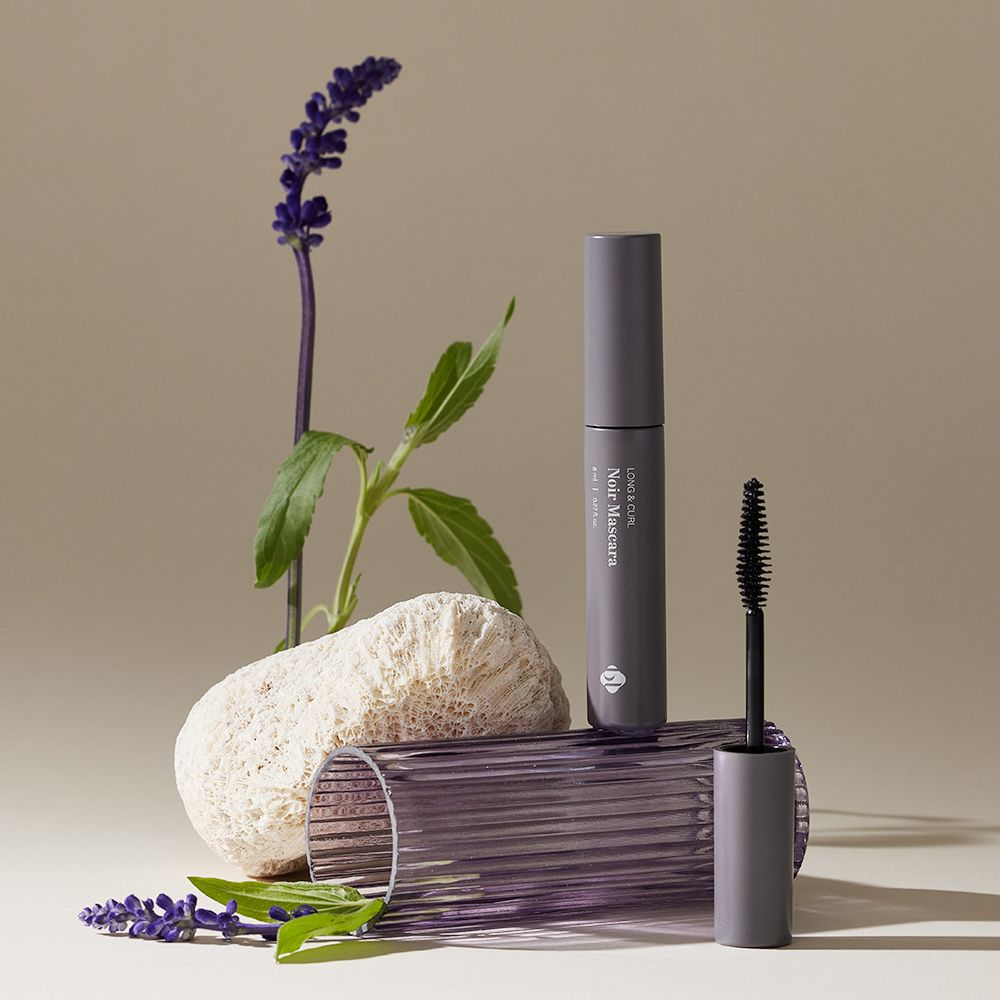 BL Lashes Noir Mascara tube and wand arranged with a purple flower, green leaves, and a textured white stone on a ribbed purple glass cylinder.