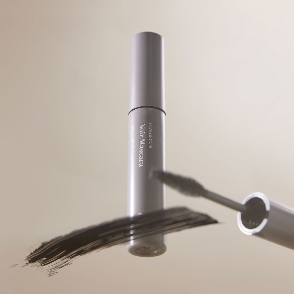 BL Lashes Noir Mascara tube standing upright, with its wand resting near a smudge of black mascara on a reflective surface.