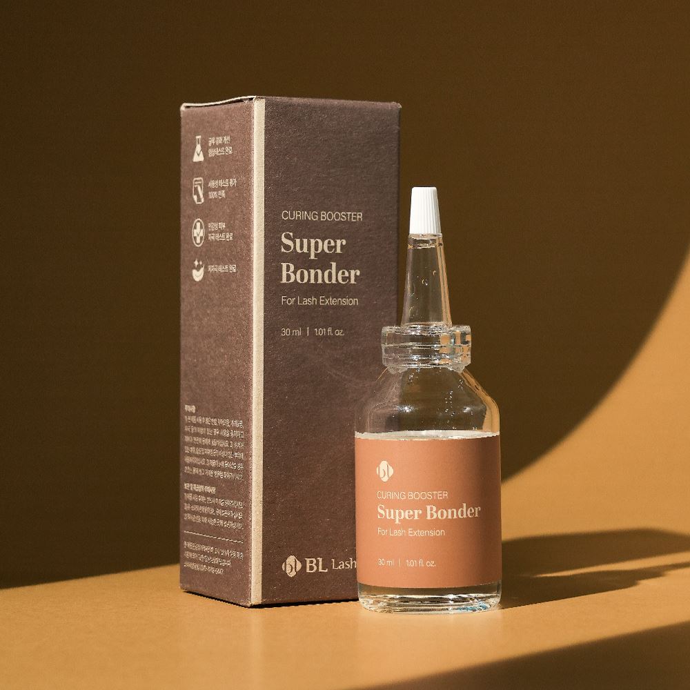 BL Lashes Super Bonder curing booster for lash extensions displayed in a clear glass bottle with a pointed applicator nozzle beside its dark brown product box on a warm-toned surface. The label reads “Curing Booster Super Bonder For Lash Extension” with the volume “30 ml / 1.01 fl. oz.” Warm directional lighting casts soft shadows, creating a professional beauty product composition that highlights adhesive curing acceleration and improved retention for eyelash extension treatments.
