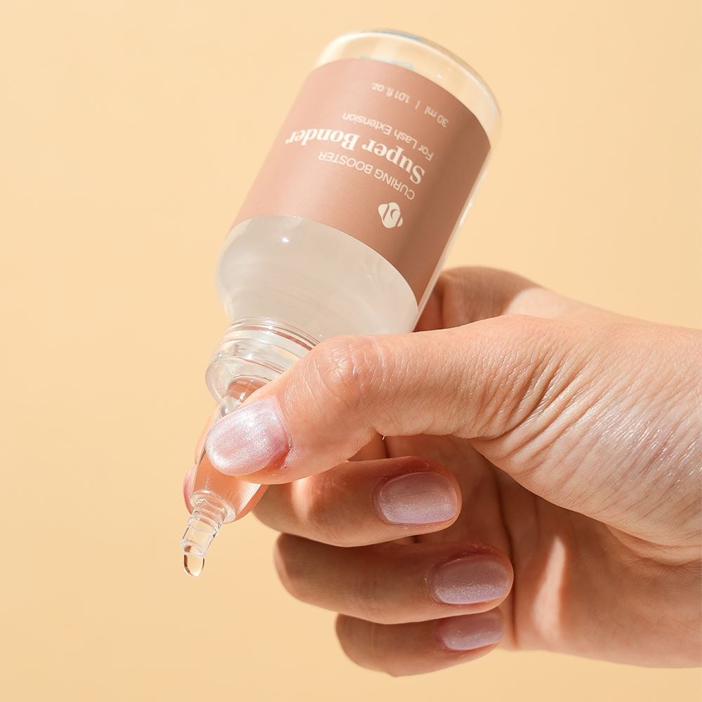 A hand holding and gently squeezing a clear glass bottle of BL Lashes Super Bonder curing booster for lash extensions with a pointed applicator nozzle. The brown label reads “Curing Booster Super Bonder For Lash Extension” with the volume “30 ml / 1.01 fl. oz.” A drop of liquid forms at the nozzle tip, demonstrating controlled dispensing. The warm beige background and soft lighting highlight precise application for accelerating adhesive curing in eyelash extension treatments.