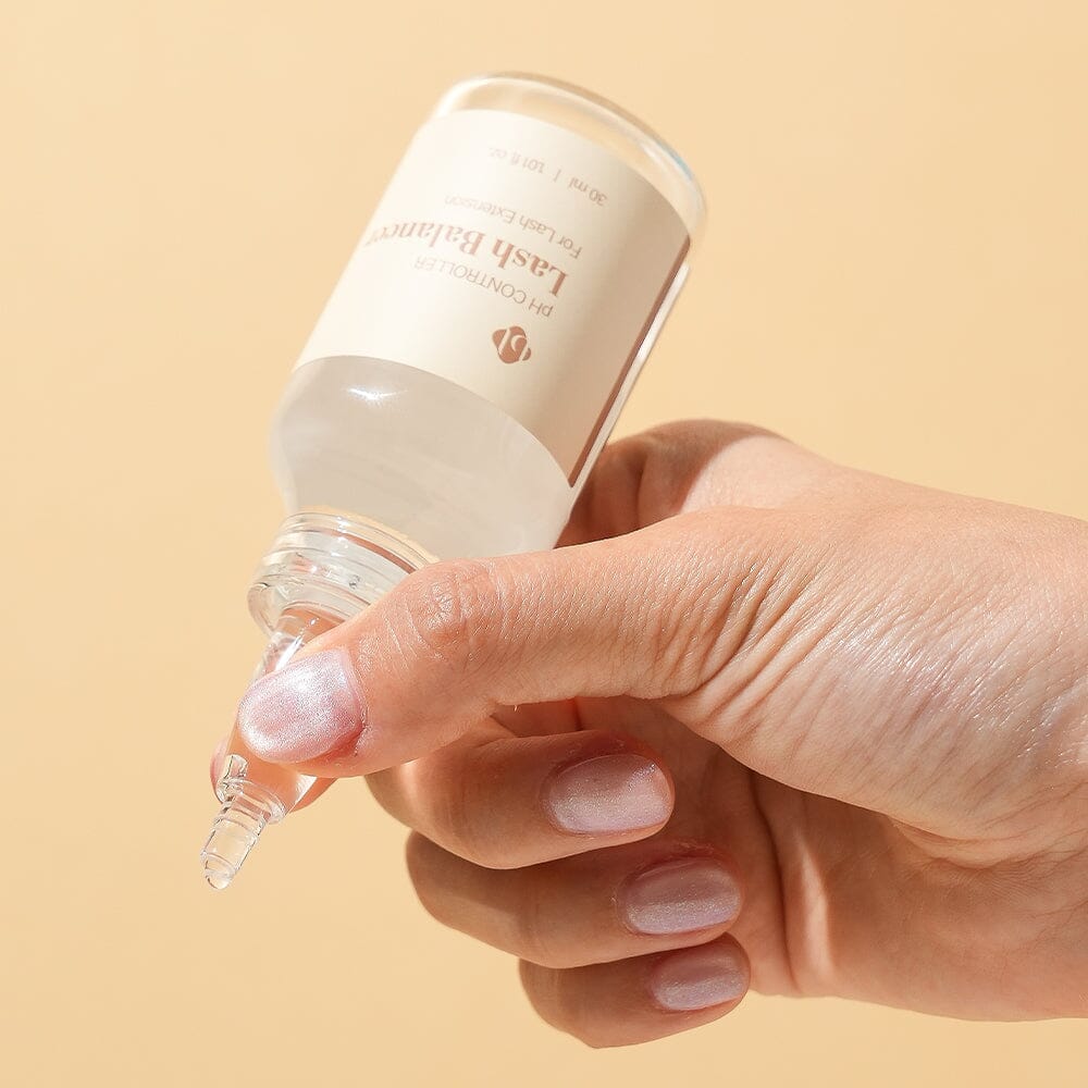 A hand holding and gently squeezing a clear glass bottle of BL Lashes Lash Balancer pH Controller for lash extensions with a pointed applicator nozzle. The beige label reads ?œpH Controller Lash Balancer For Lash Extension??and ??0 ml / 1.01 fl. oz.??A drop of liquid forms at the nozzle tip, demonstrating controlled dispensing. The warm beige background and soft lighting highlight the product?™s precision application for eyelash extension preparation and pH balancing.